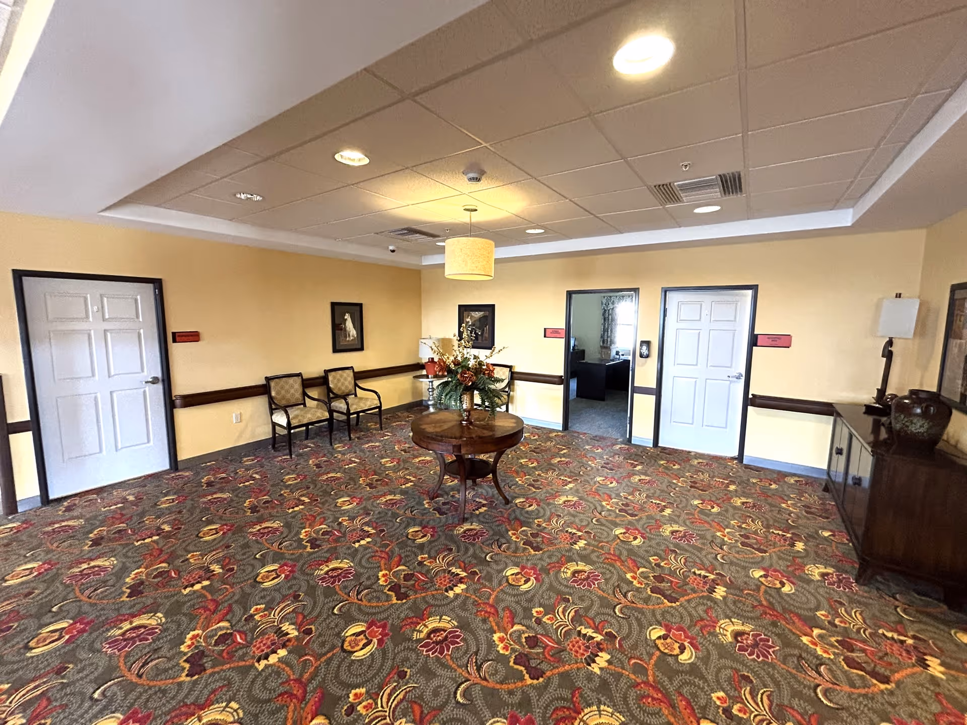 Interior view of a senior living facility common area with patterned carpet, a round wooden table with a flower arrangement in the center, two chairs against the wall, framed artwork, and three doors leading to other rooms. The walls are painted yellow and the ceiling has recessed lighting and a hanging light fixture.