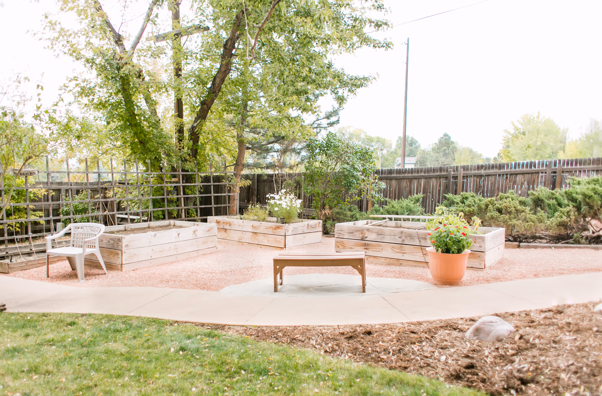 Outdoor garden area with raised wooden planter boxes containing plants and flowers, a wooden bench in the center, a white plastic chair to the left, and a large potted plant with red flowers on the right. The area is surrounded by a wooden fence and trees with green leaves.