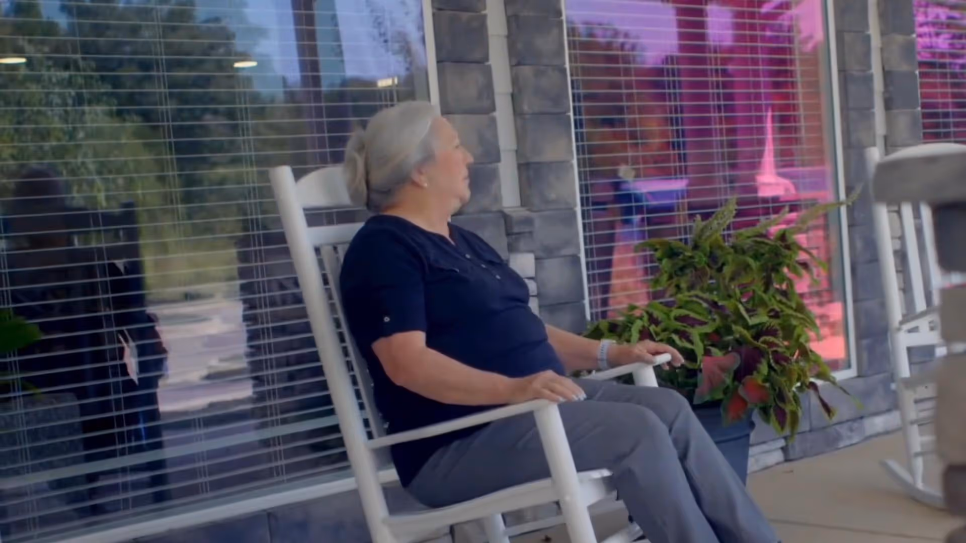 An elderly woman with gray hair tied back is sitting on a white rocking chair on a porch outside a building with large windows and stone walls. There is a potted plant with green and red leaves next to her, and the reflection of the outdoor area is visible in the windows.