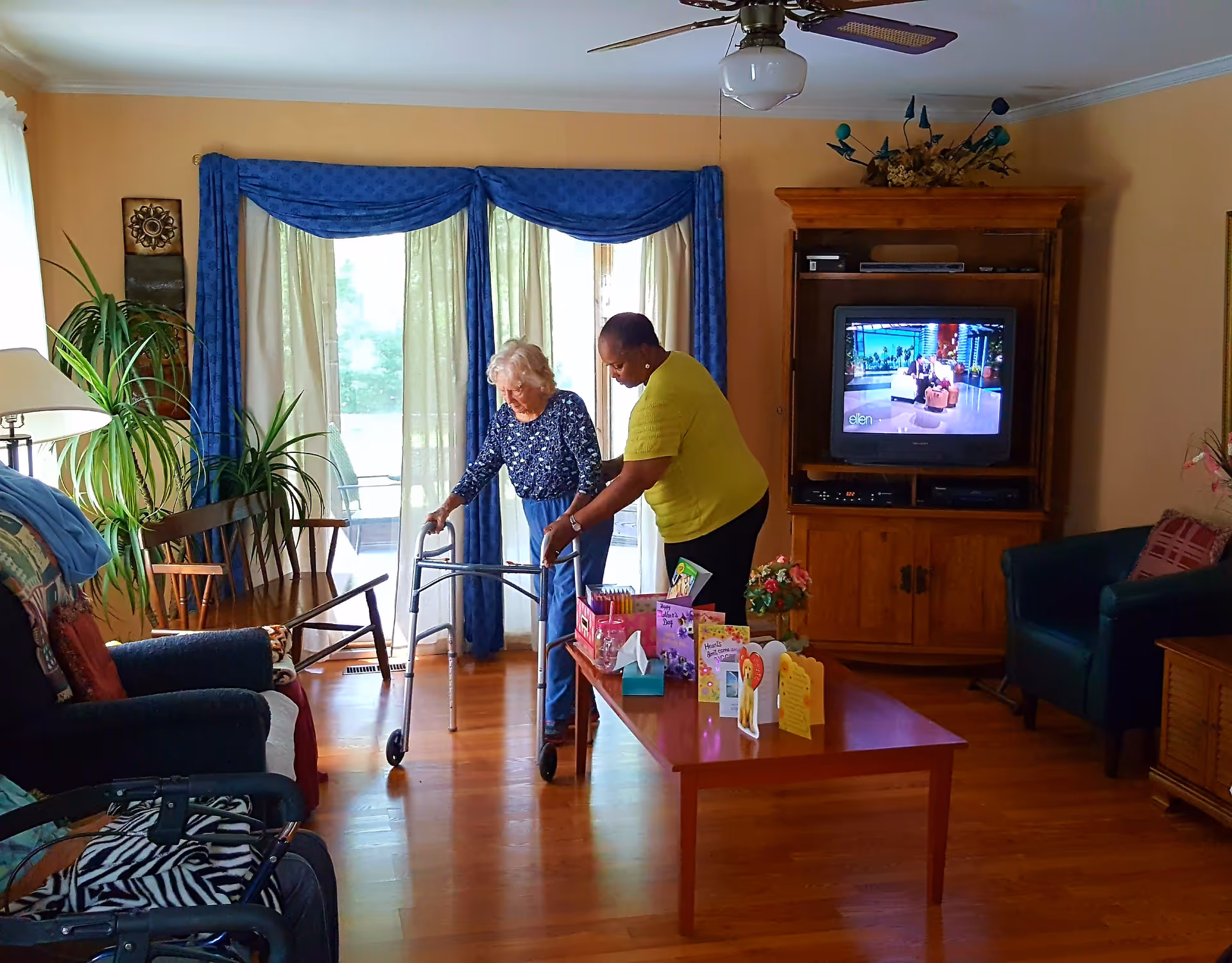 An elderly woman using a walker is assisted by a caregiver in a living room with wooden floors, blue curtains, a wooden entertainment center with a television, and a coffee table with greeting cards and flowers.