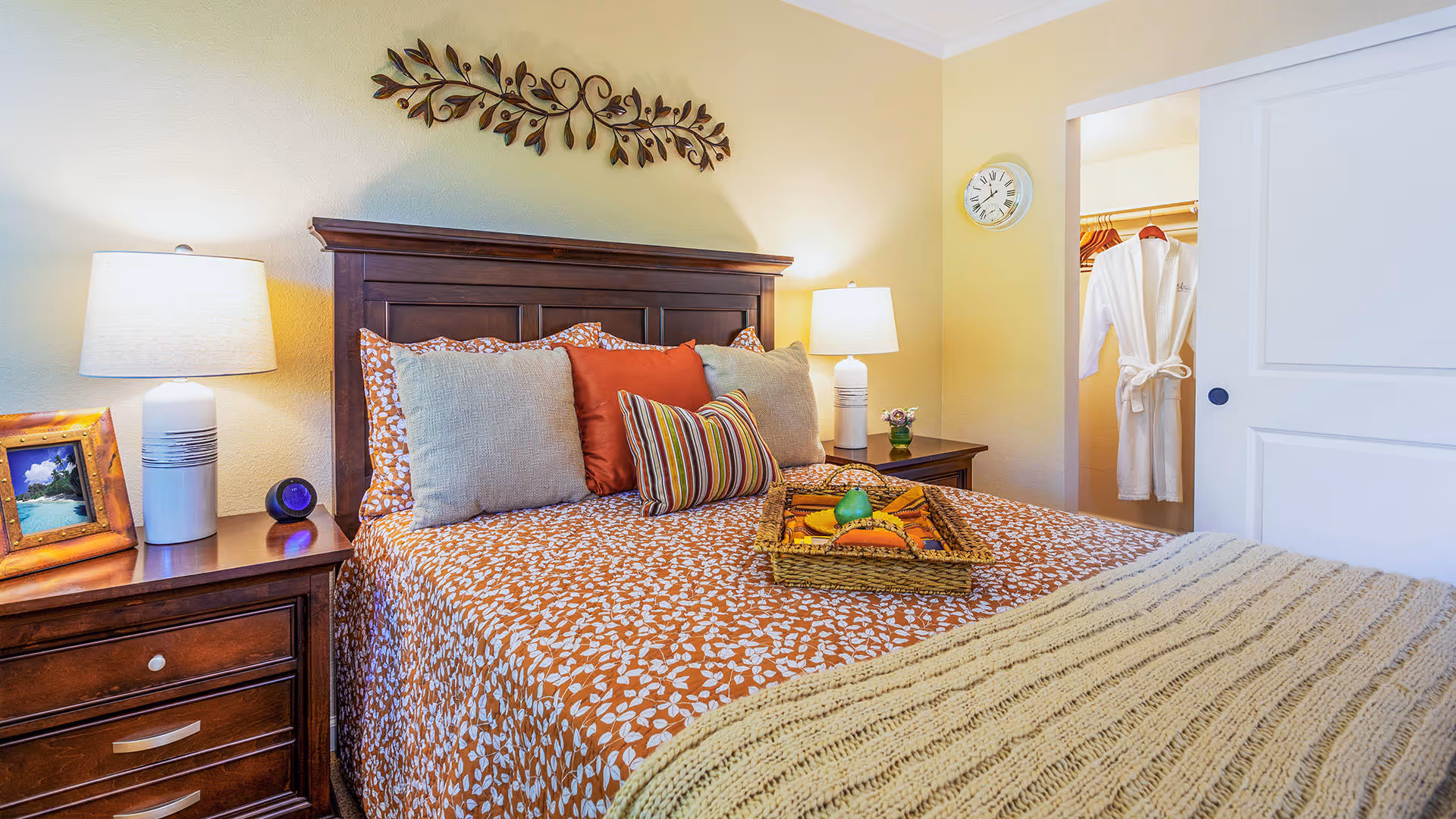 A cozy bedroom with a wooden headboard and a bed covered in an orange and white patterned bedspread, accented with multiple pillows in neutral and striped colors. There are two bedside tables with lamps, a framed photo, and a small decorative item. A woven tray with decorative fruits is placed on the bed. A white sliding door is partially open, revealing a closet with a white robe hanging inside. A decorative metal wall art piece is mounted above the headboard.