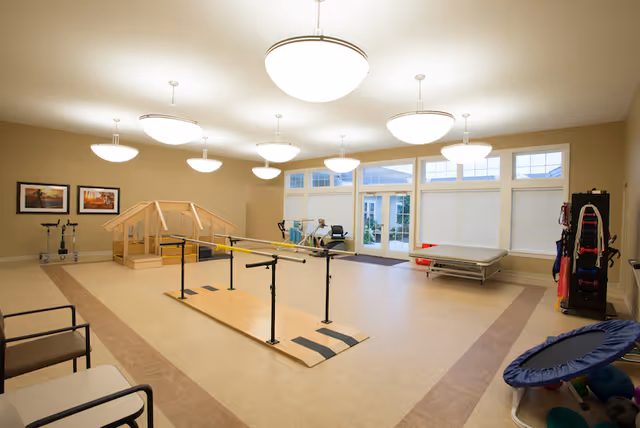 Bright rehabilitation room with parallel bars, therapy equipment, chairs and large windows.