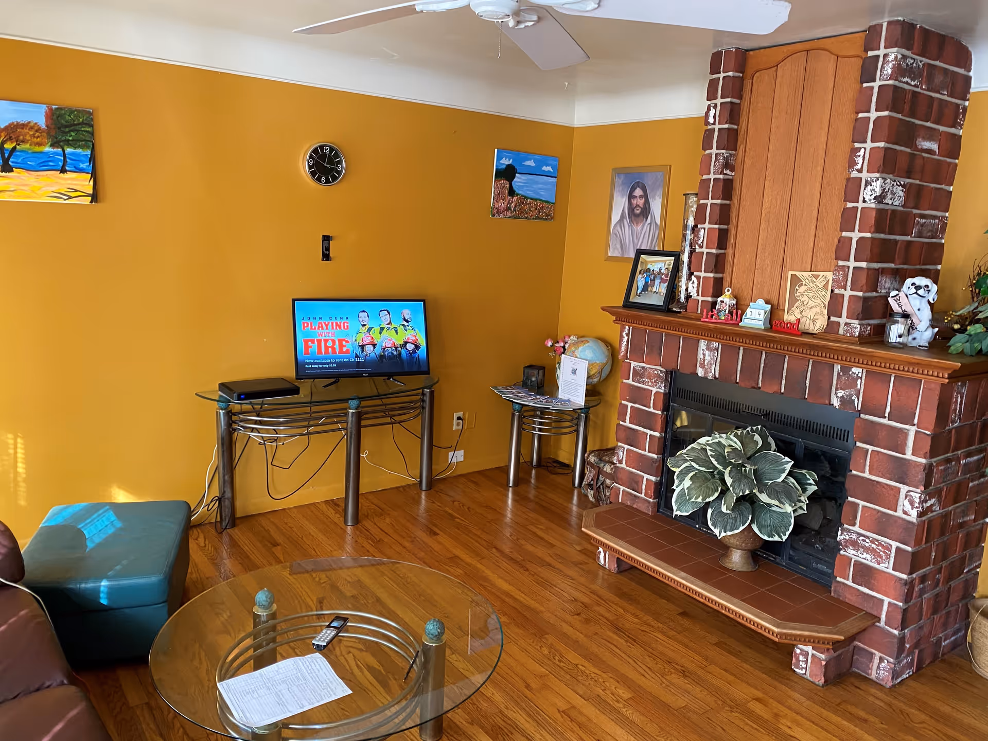 A cozy living room with mustard yellow walls, a brick fireplace decorated with framed pictures and a plant, a glass coffee table with a remote and paper on it, a small TV on a glass stand displaying a show called Playing with Fire, and colorful paintings on the walls.