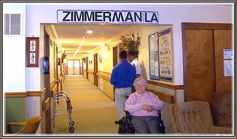 An elderly woman in a wheelchair is positioned in the foreground of a hallway in a senior living facility. Two staff members stand further down the hallway, which is well-lit and has handrails along the walls. A sign above the hallway reads 'ZIMMERMAN LA'. There is a walker near the wall and some chairs along the side.