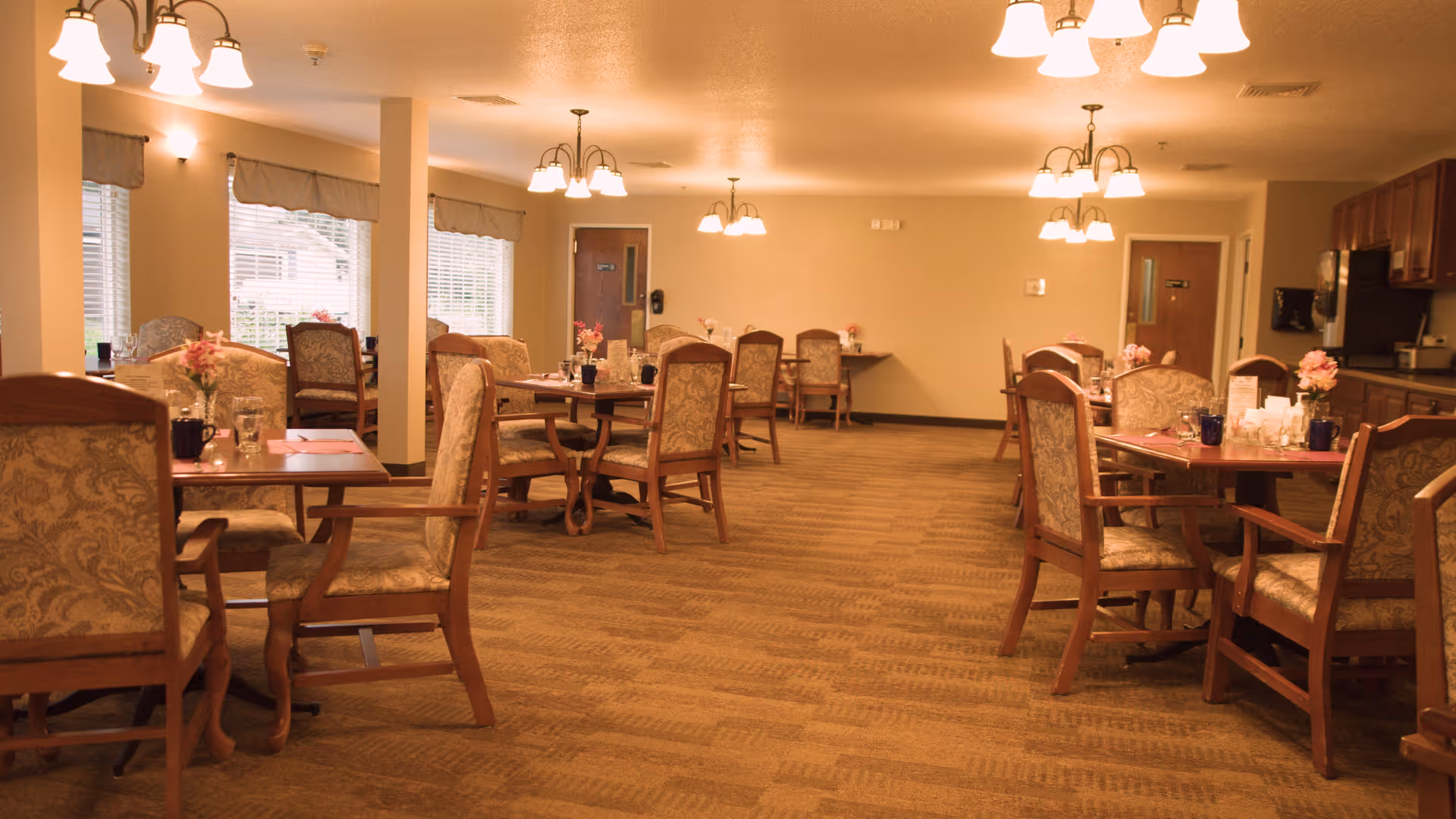 A dining room in a senior living facility with multiple wooden tables and upholstered chairs arranged neatly. Each table has small flower arrangements and place settings. The room is well-lit with ceiling lights and has large windows with blinds on the left side. There is a kitchen area visible on the right side of the room.