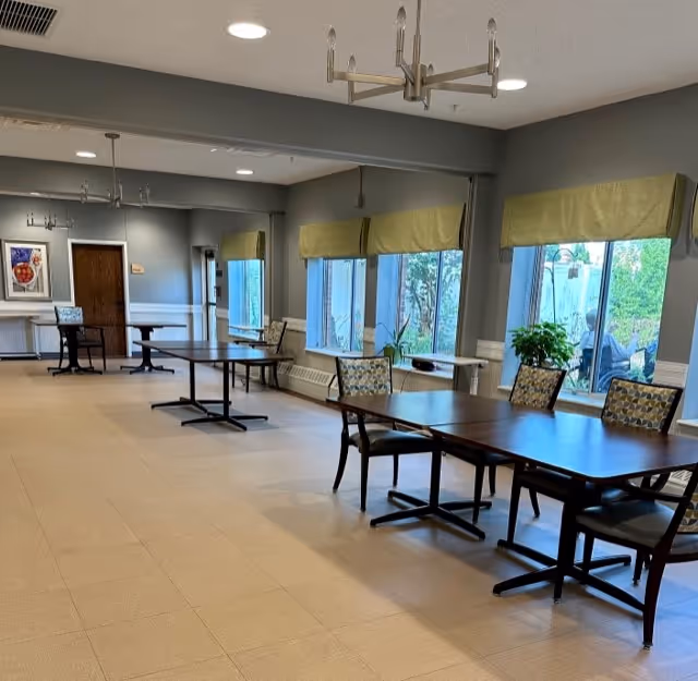 A spacious dining room with several rectangular wooden tables and cushioned chairs arranged neatly. Large windows with green valances allow natural light to fill the room, and there are plants on the windowsills. The walls are painted gray with white trim, and there is a chandelier hanging from the ceiling.