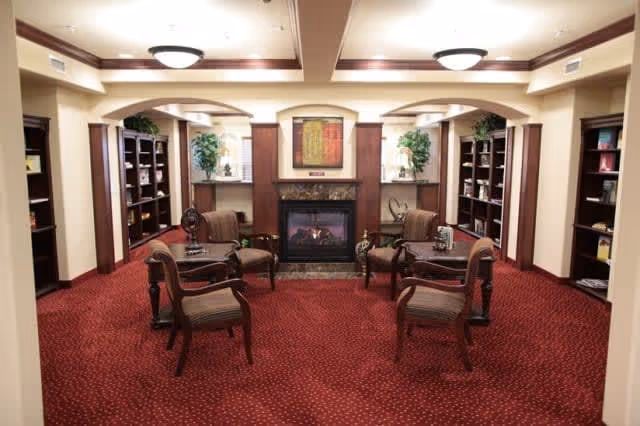 A cozy interior common area with red carpet flooring, four wooden chairs with cushions arranged around two small round tables, built-in bookshelves filled with books on either side, a fireplace with a decorative mantle in the center, and two potted plants on either side of the fireplace beneath wall-mounted lamps.