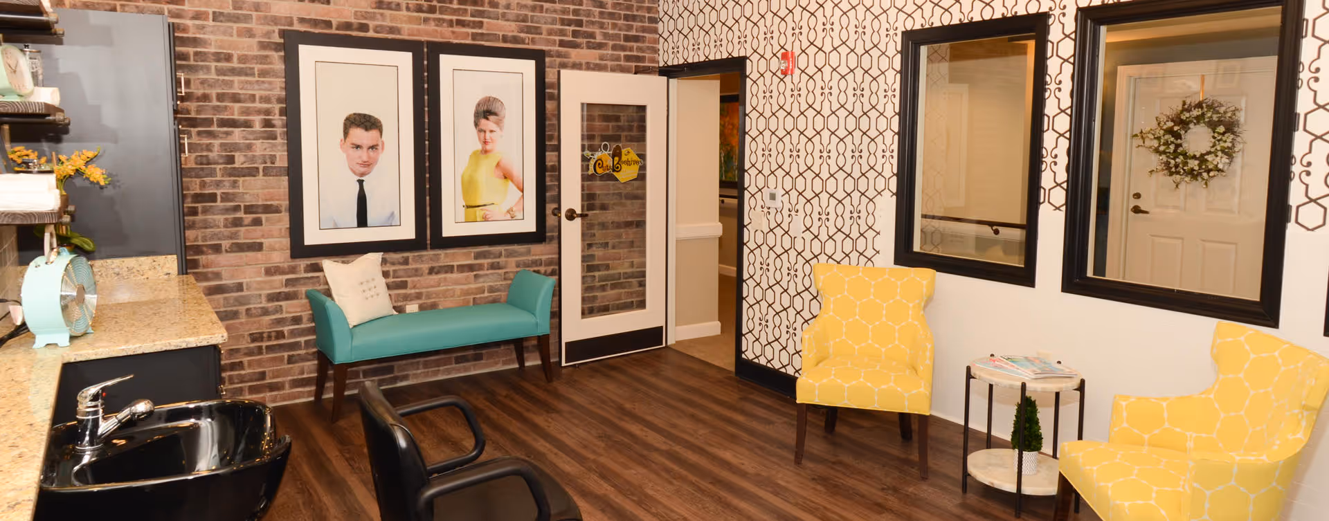 A cozy interior room with a brick accent wall featuring two large framed portraits. There is a teal bench with a white pillow beneath the portraits. The room has a wooden floor and patterned wallpaper on the adjacent wall. Two yellow armchairs with a small round table between them are positioned near two large mirrors. A door with a wreath and a glass panel is visible, along with a black sink and countertop on the left side.
