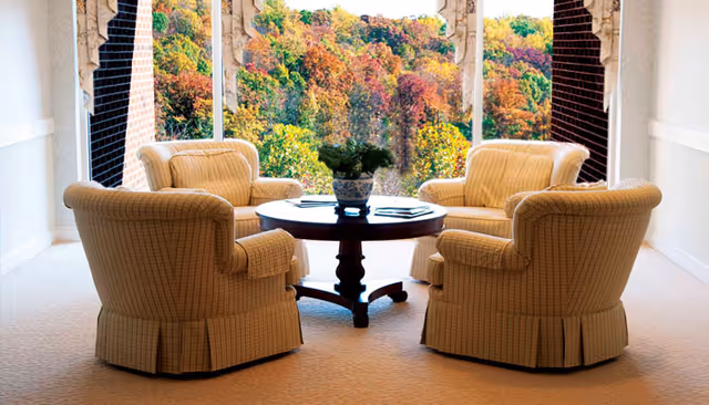 A cozy seating area with four beige upholstered armchairs arranged around a round wooden coffee table with a potted plant on top. The seating area is indoors, near large open windows that reveal a colorful autumn forest outside.