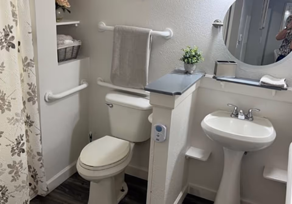 A clean bathroom featuring a white toilet with a gray towel hanging on a grab bar above it, a white pedestal sink with a faucet, a round mirror above the sink, a small potted plant on a half wall divider, and a floral shower curtain on the left side.