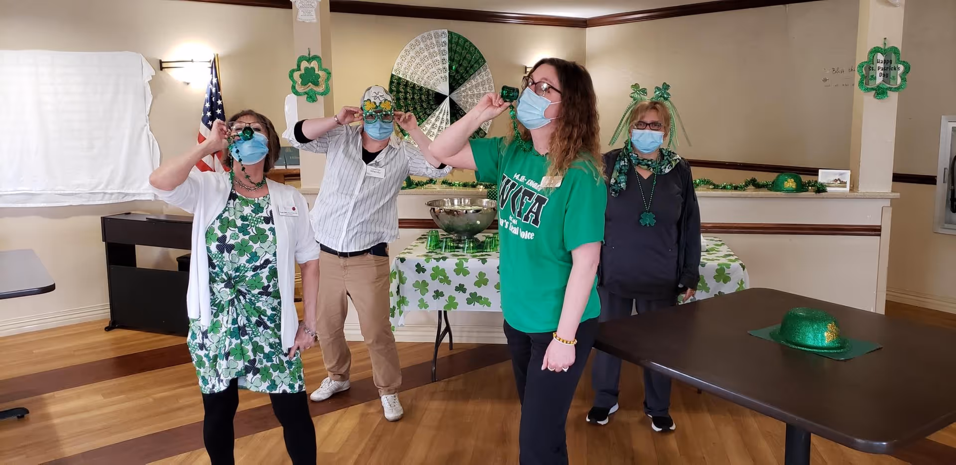 Four people wearing face masks and St. Patrick's Day themed clothing and accessories stand in a decorated room. They are holding small green cups to their mouths as if drinking. The room has shamrock decorations, a table covered with a shamrock tablecloth, and a green glittery hat on a nearby table.