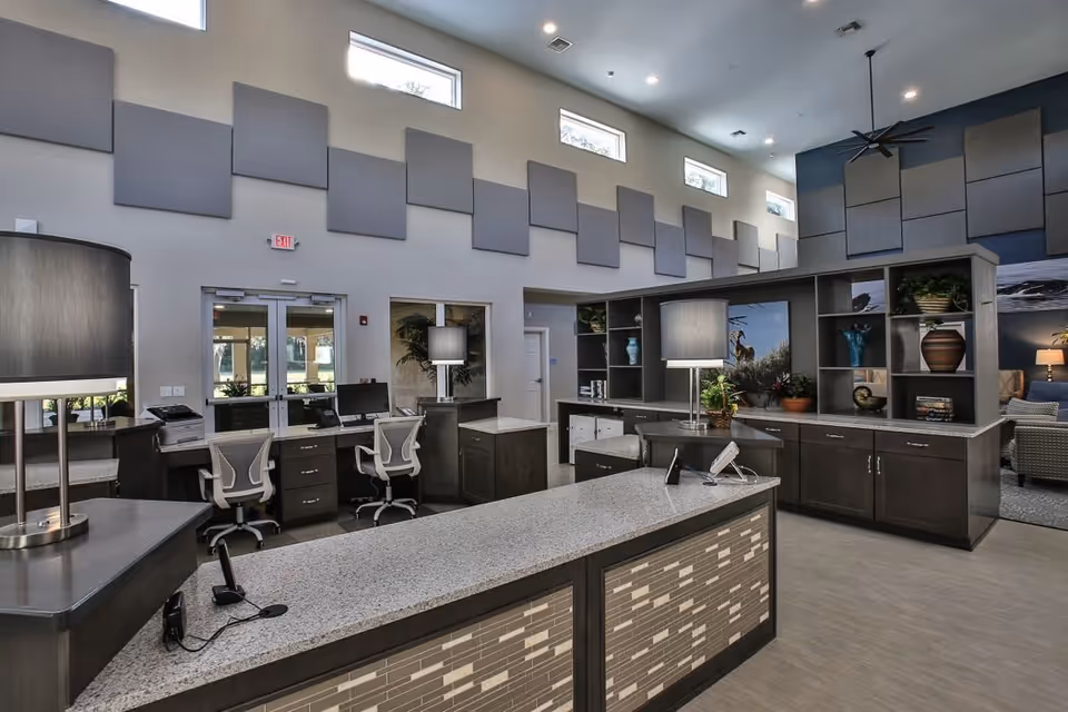Modern reception area with a long granite countertop desk, two office chairs, multiple lamps, and decorative shelving with plants and vases. The room has high ceilings with small rectangular windows near the top and sound-absorbing panels on the walls.