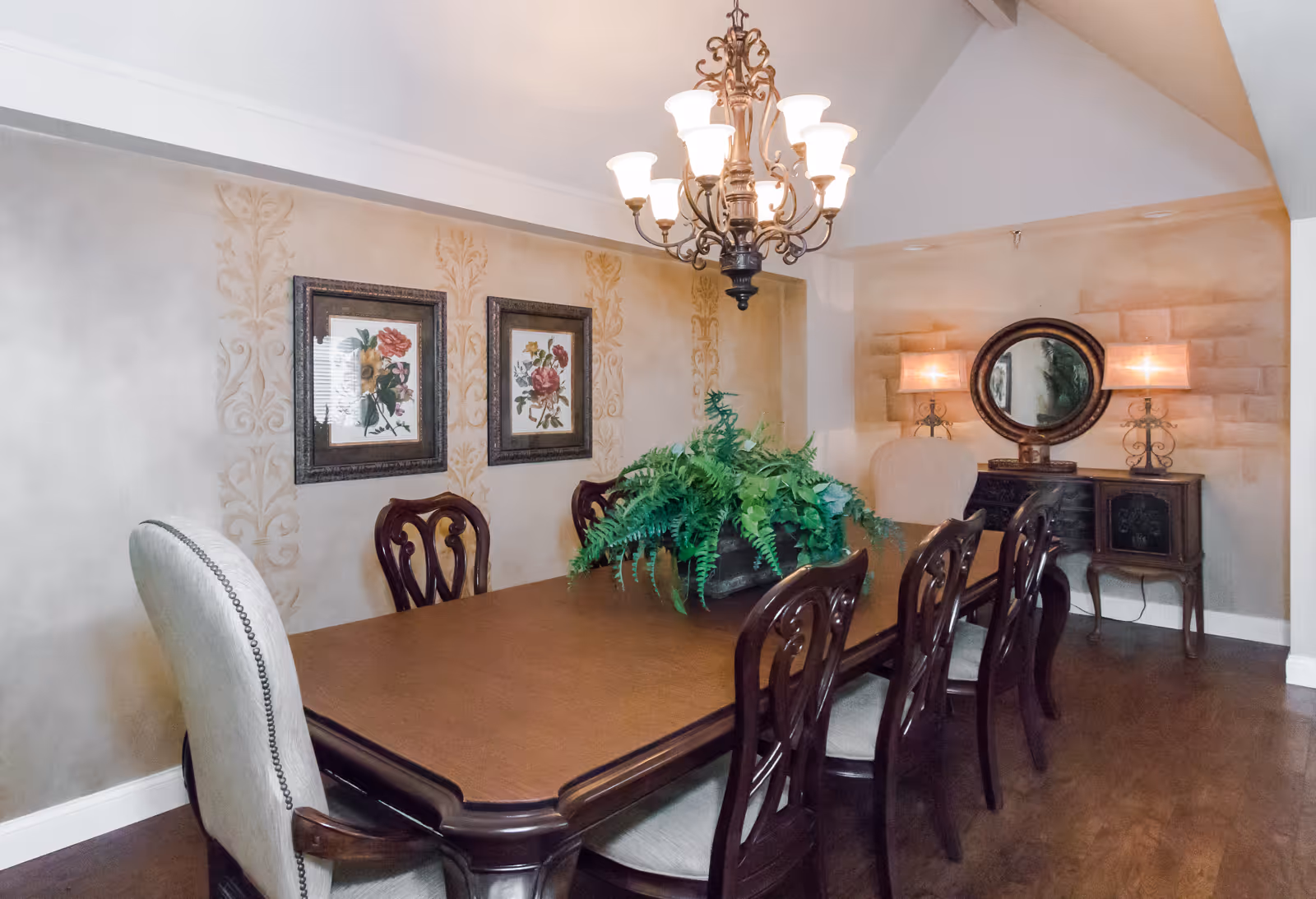 Formal dining room with a long wooden table and chairs, a chandelier, framed floral artwork, and a sideboard with mirror and lamps.