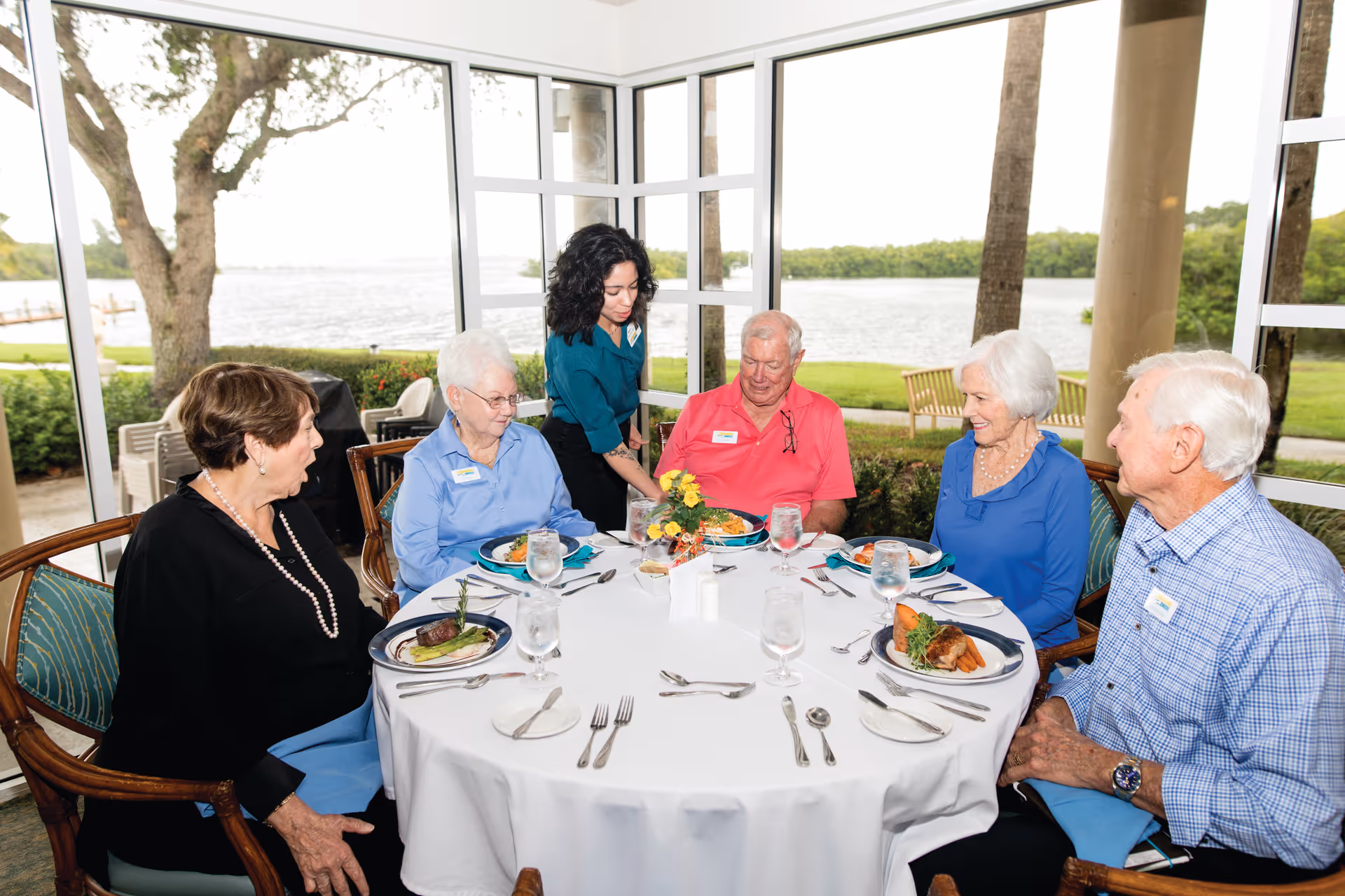 A group of older adults seated around a dining table being served by a staff member in a room with large windows overlooking a waterfront.