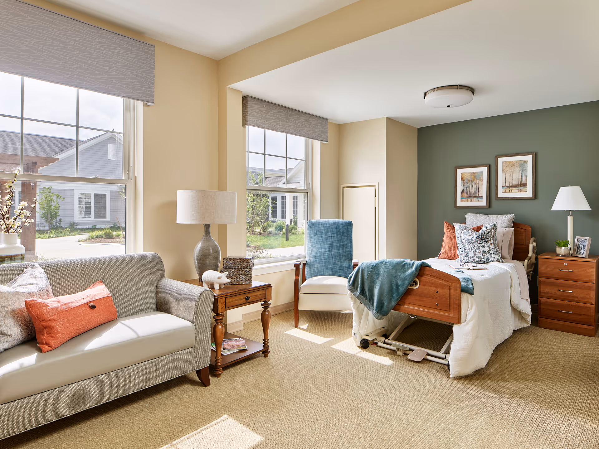 A bright and cozy bedroom in a senior living facility with a hospital-style bed, a wooden nightstand with a lamp and framed photo, a blue armchair, and a beige sofa with decorative pillows. Two large windows let in natural light, and the walls are painted in soft beige and green tones with framed artwork above the bed.