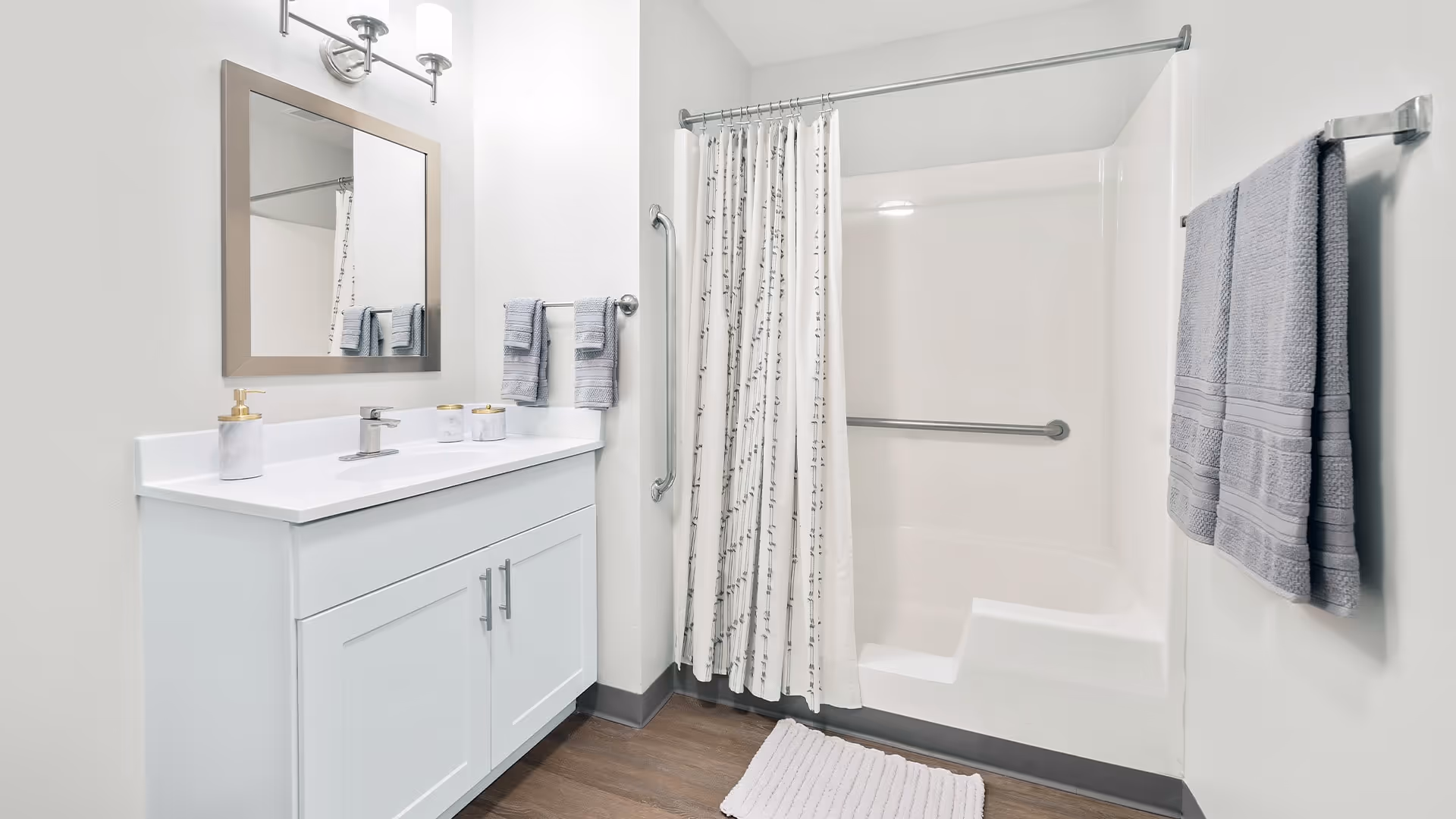 A clean and modern bathroom featuring a white vanity with a sink, a large mirror with a silver frame, and a wall-mounted light fixture above. There are two gray hand towels hanging on a towel bar next to the vanity. The bathtub has a white shower curtain with a subtle pattern, a built-in seat, and grab bars for safety. A gray towel is hanging on a towel rack on the right wall, and a white bath mat is on the wooden floor.
