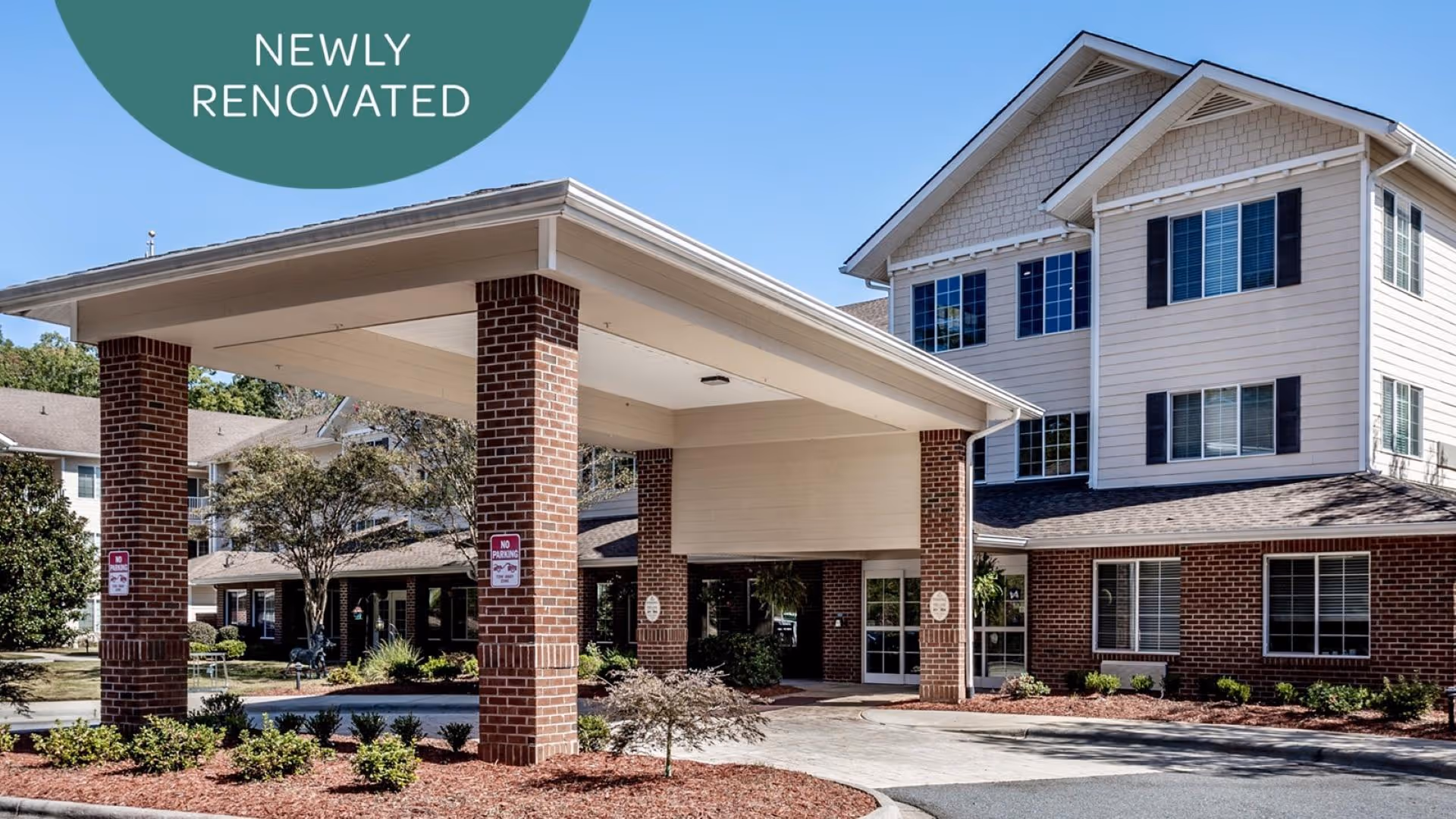 Exterior view of a newly renovated senior living facility named Holiday Willow Grove, featuring a covered entrance supported by brick pillars, landscaped bushes, and a clear blue sky.