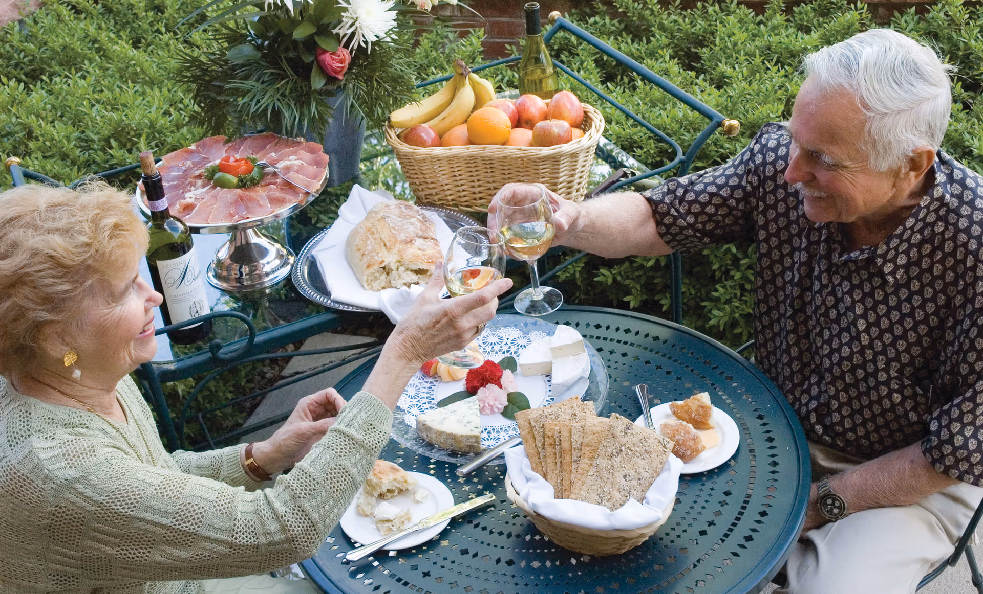 An elderly man and woman sitting at a round outdoor metal table, clinking wine glasses in a toast. The table is set with a basket of crackers, a plate of cheese, a loaf of bread, a basket of fruit, and a bottle of wine. They are surrounded by greenery and flowers.