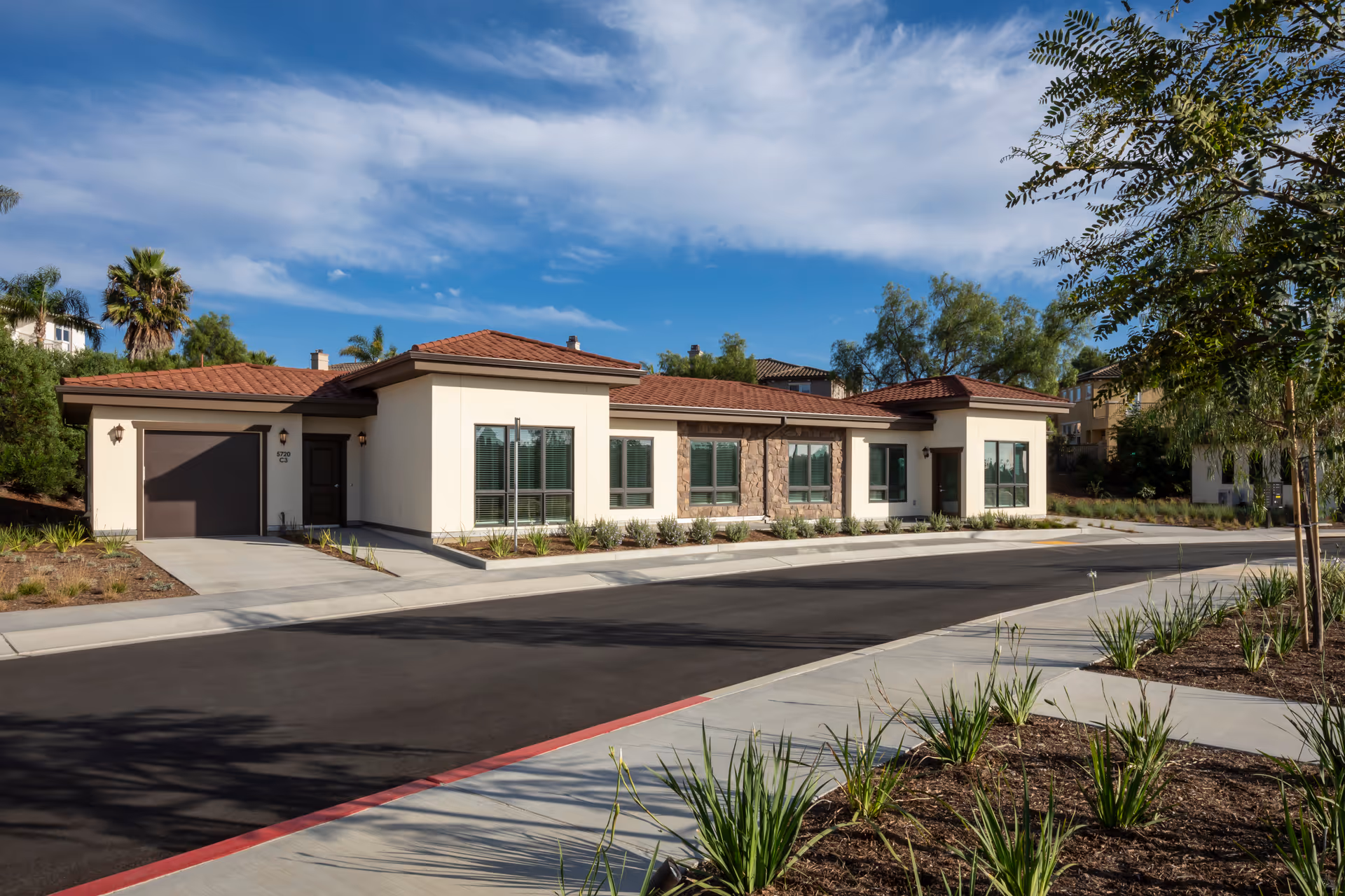 Exterior view of a single-story residential building with a tiled roof, large windows, and a garage, surrounded by landscaped plants and trees under a partly cloudy blue sky.