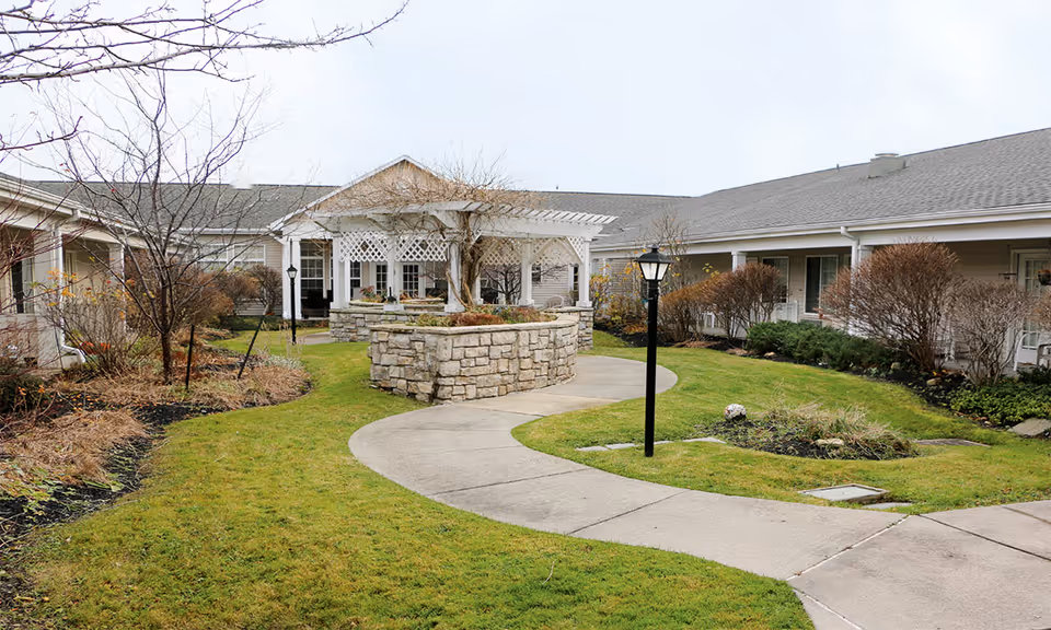 Landscaped courtyard with a curved concrete walkway, stone planter, pergola and lamp post surrounded by single-story buildings.