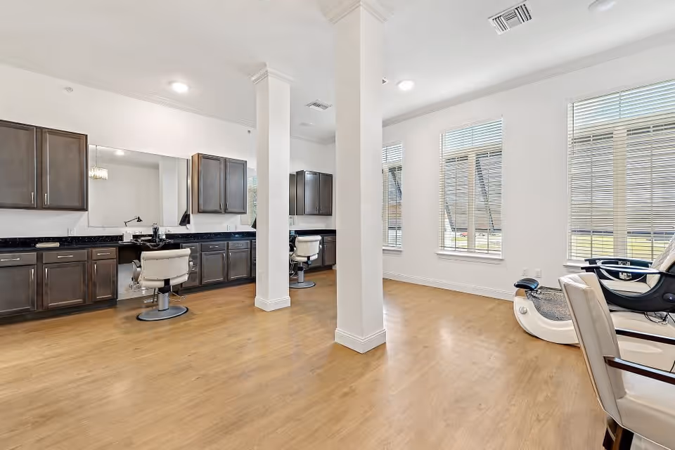 Interior view of a salon area in a senior living facility with wooden flooring, white walls, large windows with blinds, two styling chairs in front of a long counter with mirrors and cabinets, and a foot spa chair on the right side.