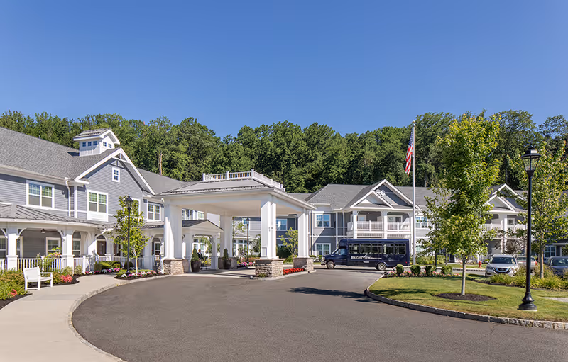 Exterior view of Brightview Warren Senior Assisted Living & Memory Care facility on a clear sunny day. The building is two stories with gray siding and white trim. There is a covered entrance with white pillars and stone bases. A small shuttle bus and several cars are parked near the entrance. Trees and landscaped greenery surround the driveway and building.