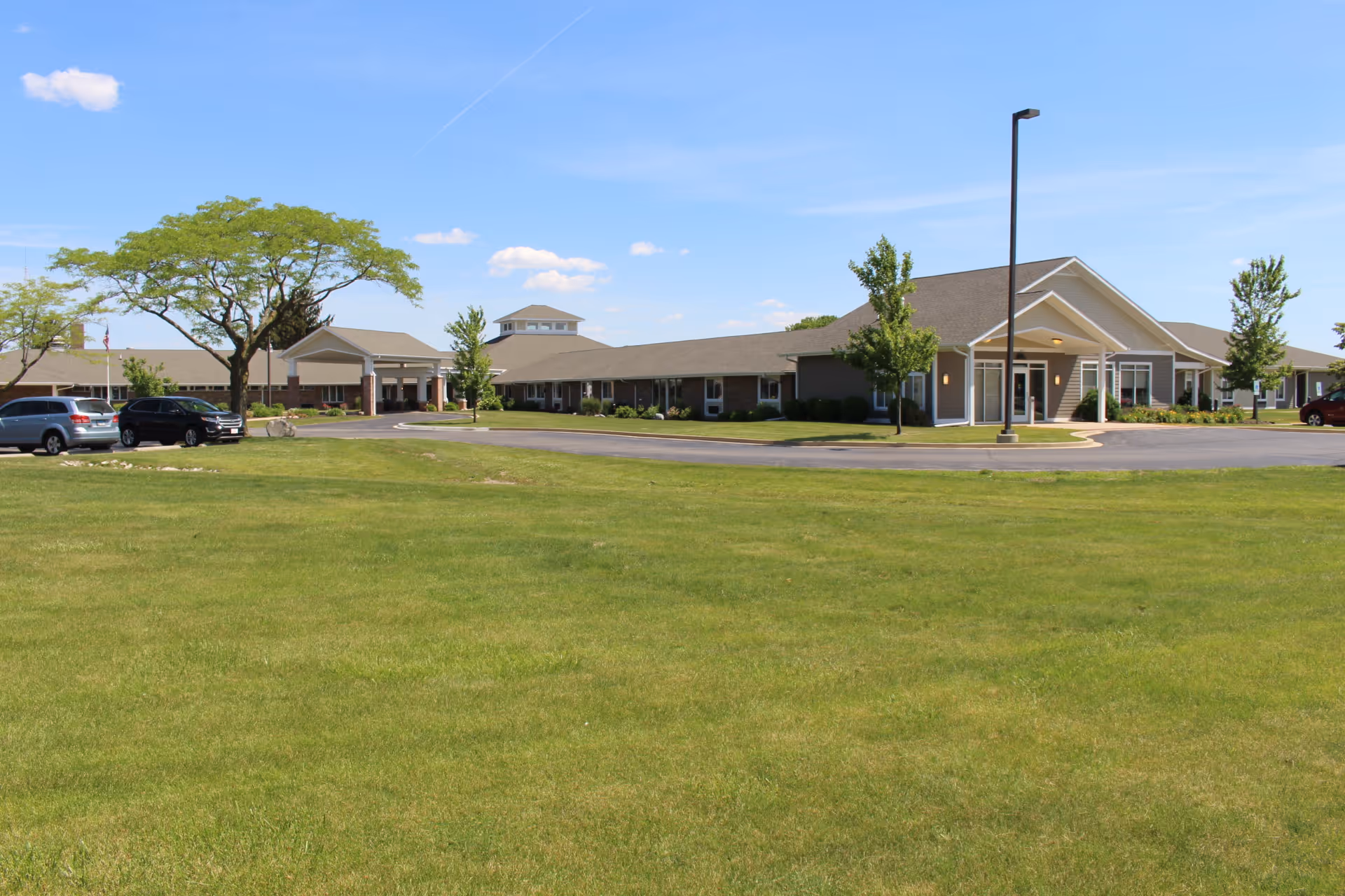 One-story building with a covered entrance, circular driveway, parked cars and a wide grassy lawn under a blue sky.
