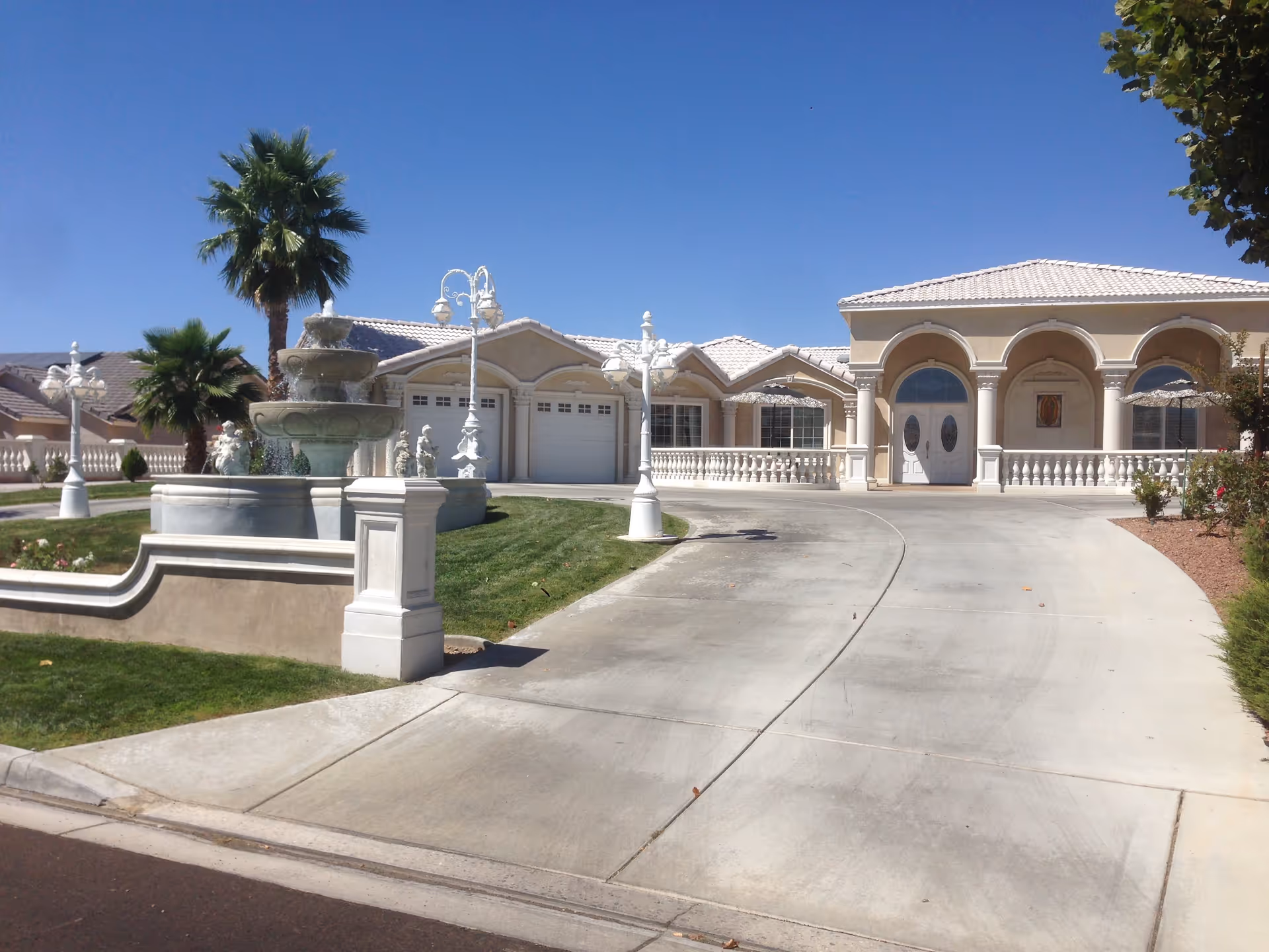 Exterior view of a large, elegant single-story building with a white tiled roof, arched windows, and columns at the entrance. A wide concrete driveway leads up to the building, and there is a multi-tiered stone fountain surrounded by green grass and palm trees in front. The sky is clear and blue.