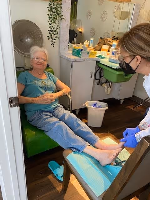 An elderly woman is seated comfortably in a green chair with her feet resting on a footrest covered with a blue disposable sheet. A caregiver wearing a black face mask and blue gloves is attending to the woman's feet in a room with a sink, counter, and various supplies. The setting appears to be a personal care or medical treatment area.