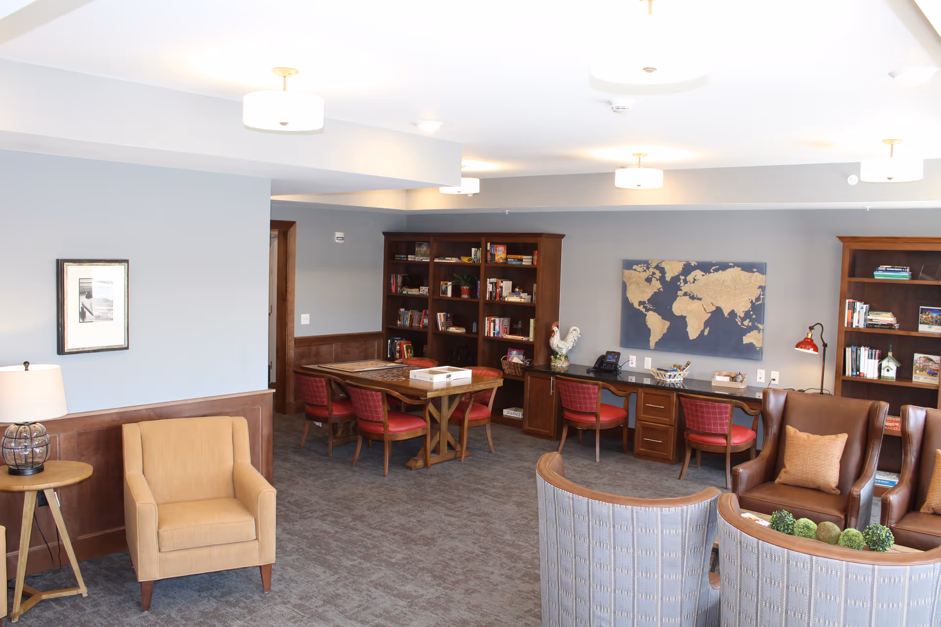A cozy common area with comfortable seating including armchairs and a curved sofa, a wooden table with red cushioned chairs, bookshelves filled with books, a world map on the wall, and soft lighting from ceiling fixtures and a table lamp.
