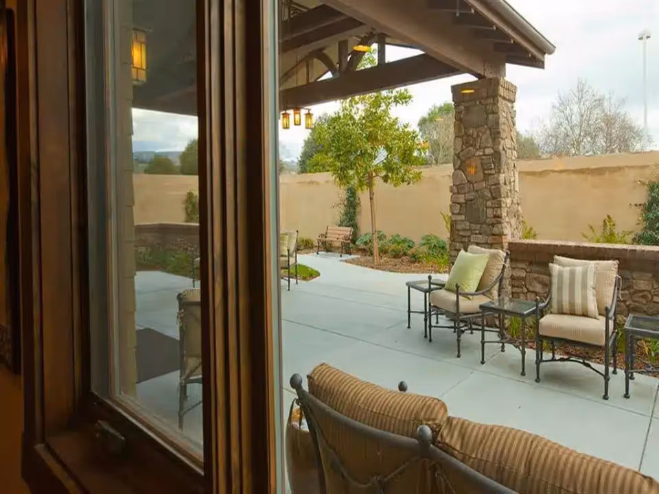 View through a window showing an outdoor patio area with cushioned chairs, small tables, stone pillars, a tree, and a beige wall surrounding the space under a covered roof.