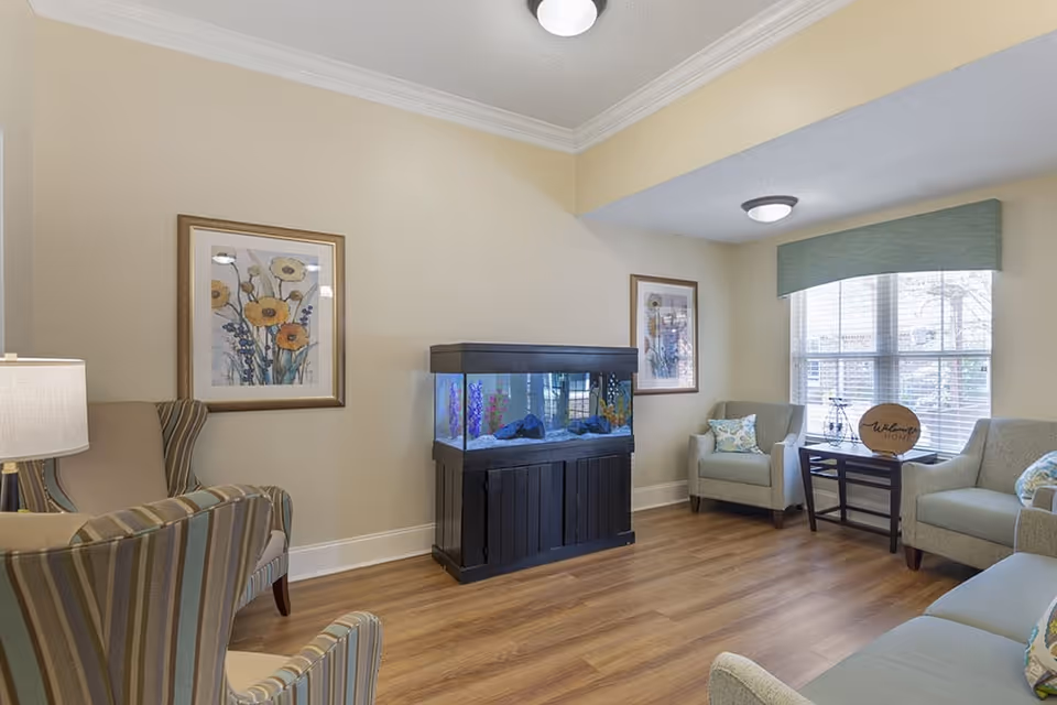 A cozy living room with light beige walls and wood flooring. The room features a large fish tank on a black stand against the wall, two framed floral paintings, a window with a green valance, and several upholstered chairs and a sofa arranged around a small dark wooden table with a decorative 'Welcome Home' sign.