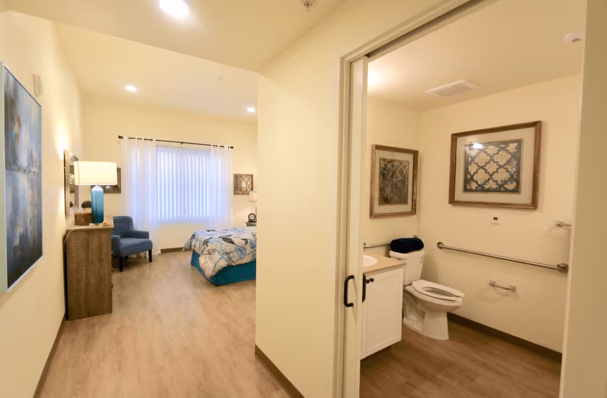 View of a senior living facility room showing a bedroom area with a bed, blue chair, dresser with a lamp, and artwork on the walls. Adjacent to the bedroom is a bathroom with a toilet, sink, grab bars, and framed artwork on the walls.