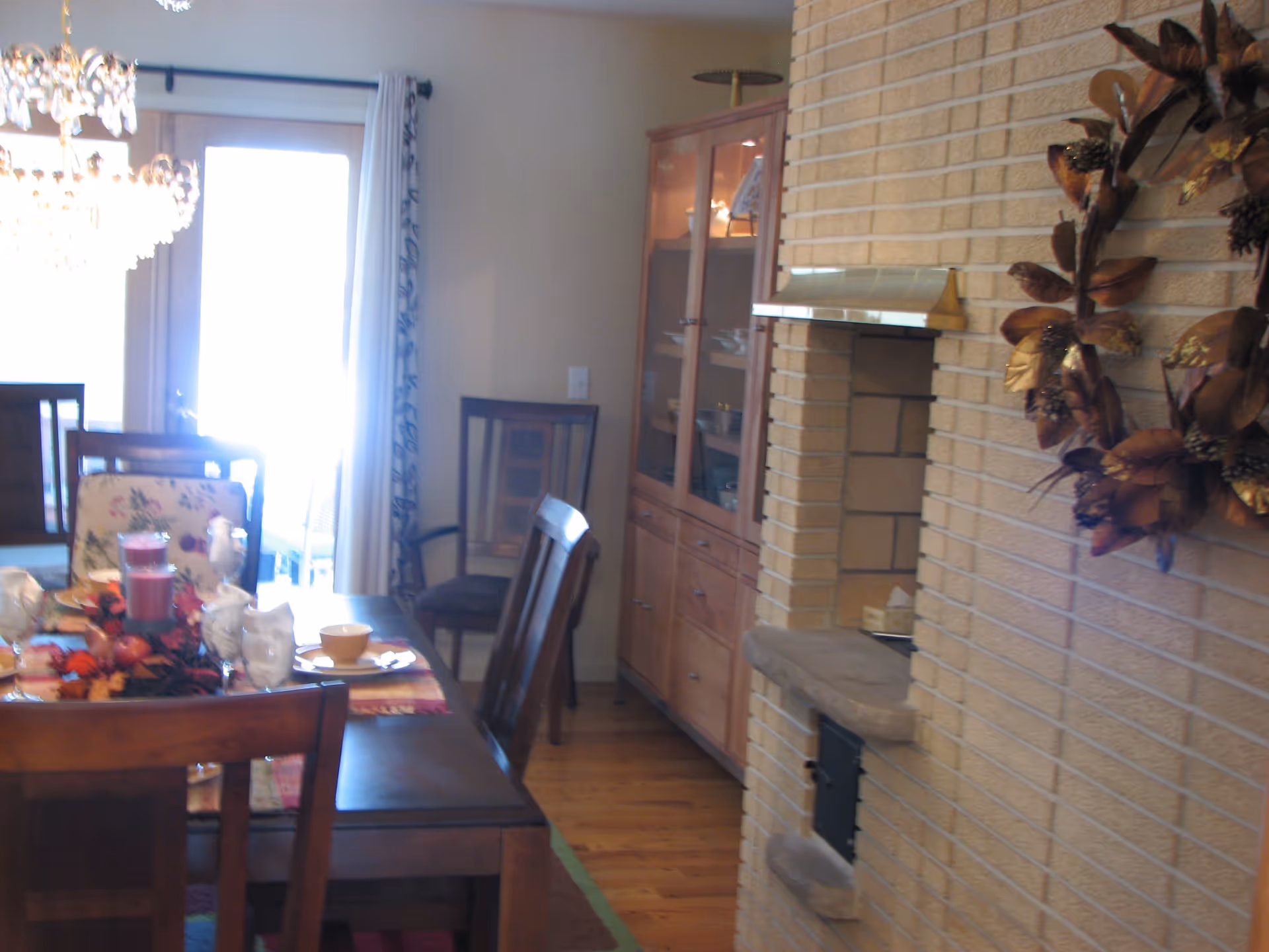 Dining room with a wooden table set for a meal, including plates, bowls, glasses, and a centerpiece with a candle and autumn leaves. There are wooden chairs around the table, a glass-front cabinet with dishes, a brick fireplace with a decorative wreath, and a glass door with curtains letting in bright light.