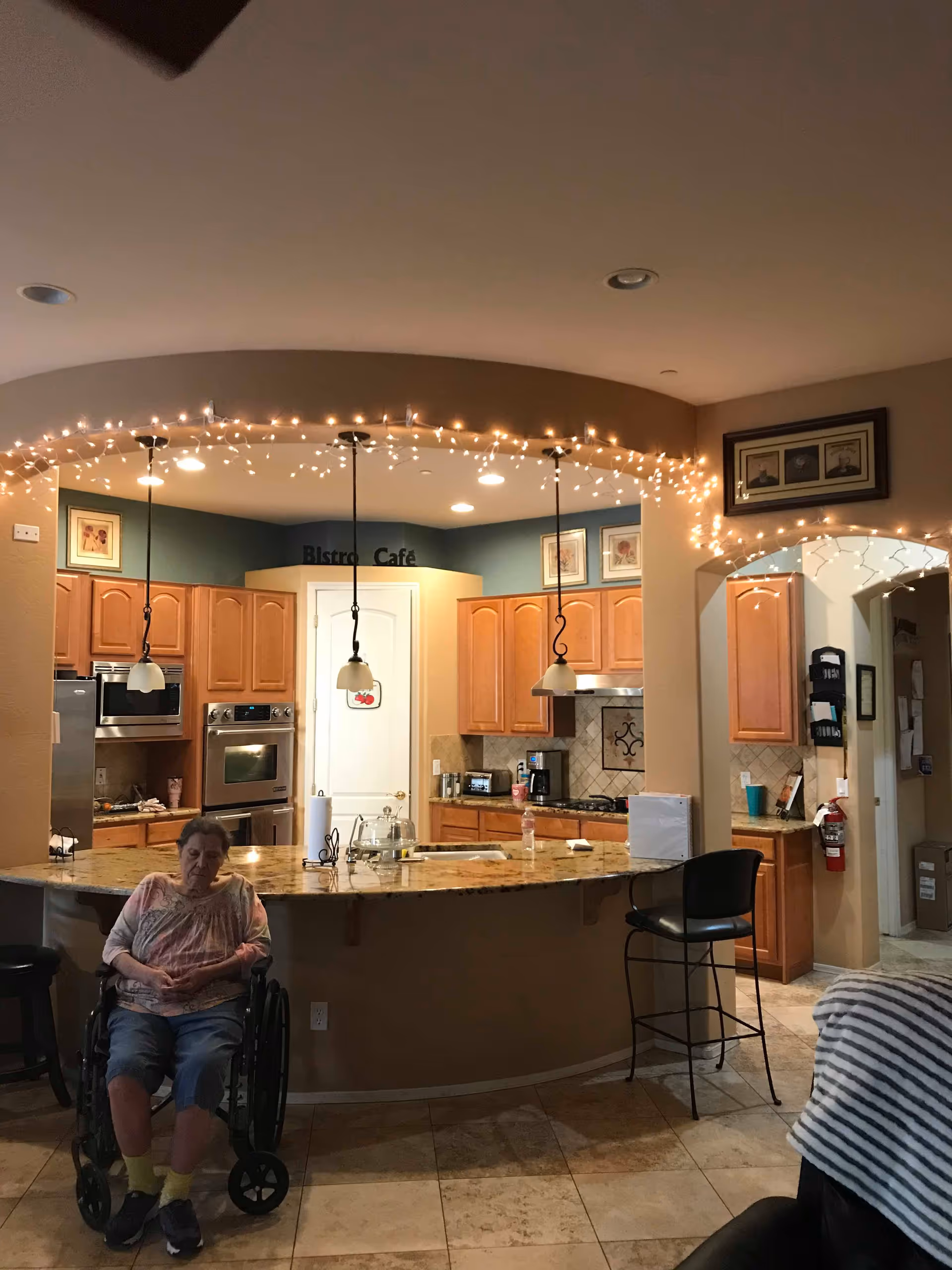 Interior view of a senior living facility kitchen area with wooden cabinets, granite countertops, and hanging pendant lights. A string of small white fairy lights decorates the archway above the kitchen counter. An elderly woman in a wheelchair is seated in front of the counter. The kitchen has a refrigerator, oven, microwave, and coffee maker. There are framed pictures on the walls and a fire extinguisher mounted near an archway leading to another room.