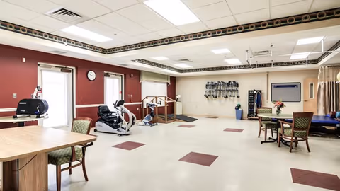 Spacious rehabilitation/activity room with exercise machines, tables and chairs, and open floor space.