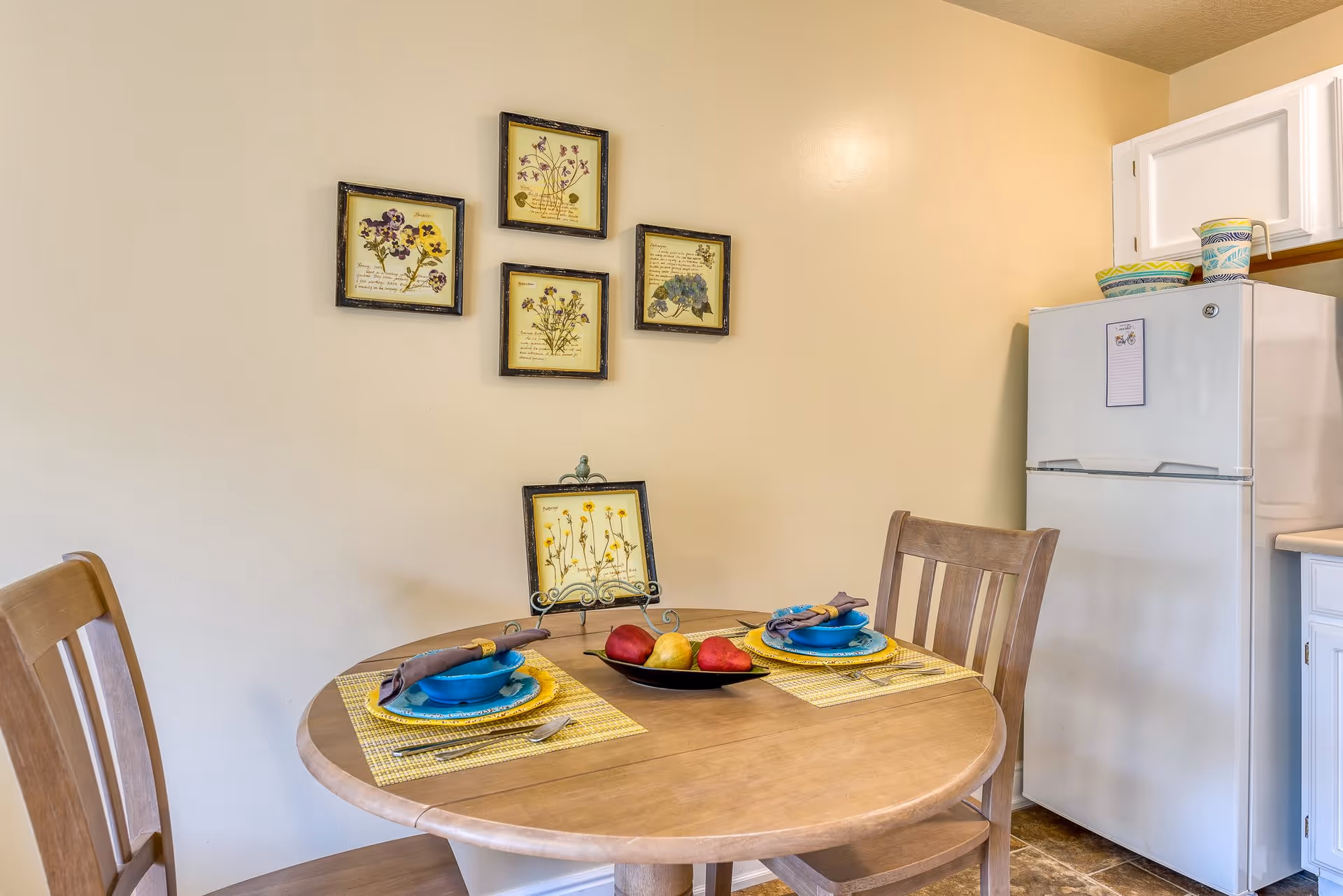 A small dining area with a round wooden table set for two with blue bowls, yellow placemats, and brown napkins. A black dish with two red pears and one yellow pear is in the center of the table. Behind the table, there are five framed botanical prints on a beige wall. To the right, a white refrigerator with colorful bowls and a pitcher on top is visible, along with white kitchen cabinets.
