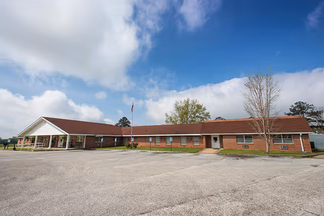 Single-story brick senior care building with a covered entrance, flagpole, and a large empty parking lot under a blue sky.