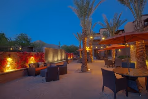 Outdoor patio area at dusk with seating arrangements including chairs and tables with orange umbrellas, palm trees, illuminated wall with red foliage, and a building in the background under a clear blue sky.