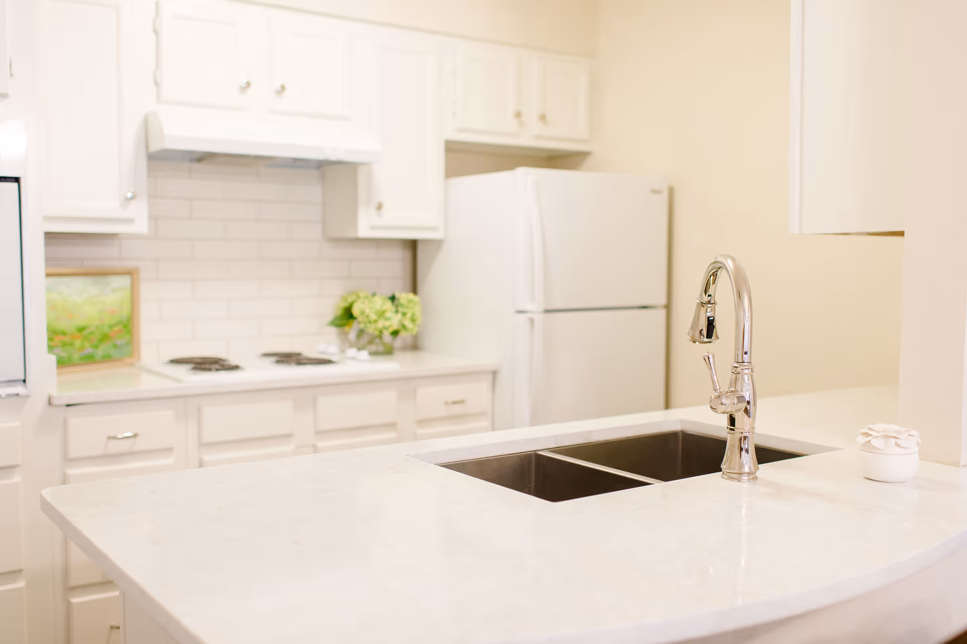 Bright white kitchen featuring a marble countertop with a double sink and chrome faucet, stove and refrigerator in the background.