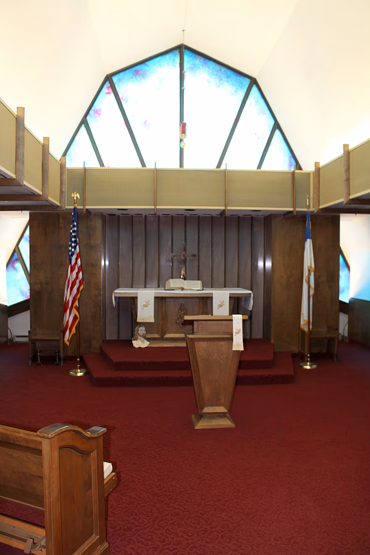 Interior view of a chapel with a wooden podium in the center, an altar with a Bible and cloths, an American flag on the left, and a Christian flag on the right. The background features stained glass windows with blue and purple hues and a wooden panel wall with a cross.