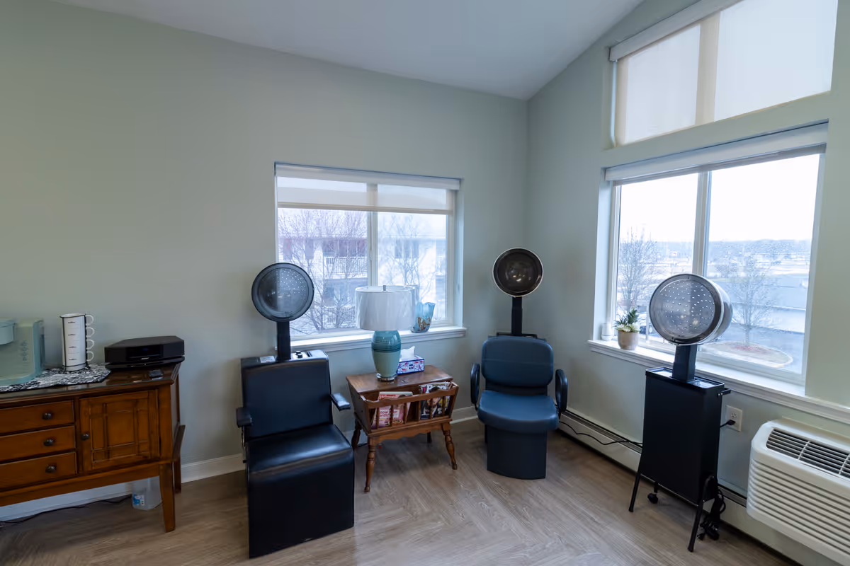 A small room with two black salon chairs equipped with hair dryers, a wooden side table with a lamp and magazines, a wooden cabinet with a microwave and coffee maker, and large windows showing an outdoor view.