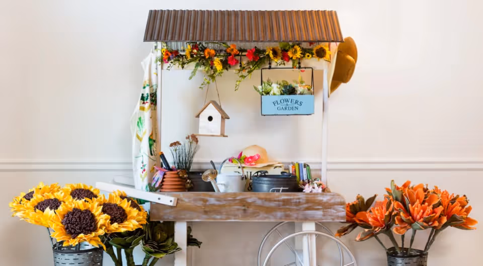 A decorative wooden garden cart with a corrugated metal roof, adorned with artificial sunflowers and orange flowers. The cart holds gardening tools, a watering can, a birdhouse, a hat, and a small planter box labeled 'FLOWERS GARDEN'. The background is a plain white wall with a chair partially visible behind the cart.