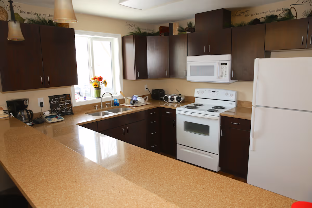 Bright residential-style kitchen with dark wood cabinets, white appliances, a sink under a window and a long tan countertop.
