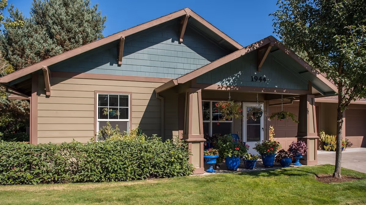 Exterior view of a single-story residential building with beige and blue siding, a covered porch with hanging flower pots, various potted plants, a tree on the right, and a well-maintained lawn in front. The building number 1944 is visible above the porch.