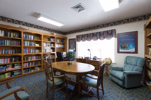 A cozy library or reading room with wooden bookshelves filled with books along the walls, a round wooden table with four chairs in the center, a comfortable blue recliner chair near a window with patterned valance curtains, a globe on a stand, and framed pictures on the walls. The room has a carpeted floor with a floral pattern and fluorescent ceiling lights.