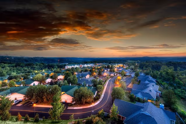 Aerial view of Longwood At Oakmont Retirement Community at dusk, showing multiple residential buildings surrounded by trees and greenery, with streetlights illuminating the roads and a dramatic cloudy sky overhead.