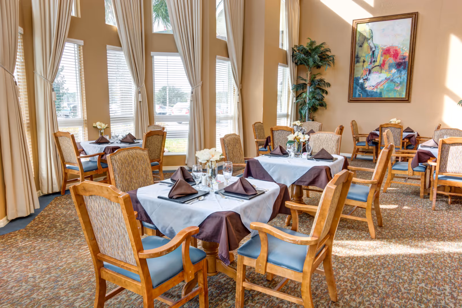 A bright dining room with several wooden tables and chairs arranged neatly. Each table is covered with a white and brown tablecloth and set with folded brown napkins, glasses, and silverware. Large windows with beige curtains allow natural light to fill the room. A colorful abstract painting and a tall green plant decorate the walls.