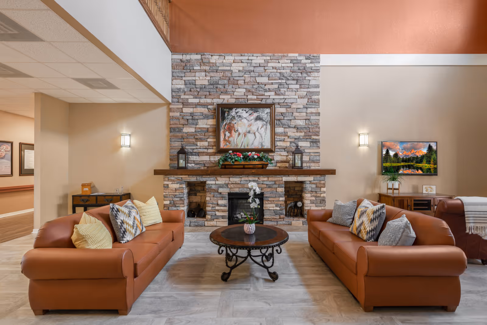 A cozy living room area with two brown leather sofas facing each other across a round wooden coffee table with a small plant. The background features a stone fireplace with a wooden mantle decorated with lanterns and a flower arrangement. Above the fireplace is a framed painting of horses. On the right wall, there is a flat-screen TV mounted above a wooden cabinet with a plant and a small picture frame. The room has warm lighting and a neutral color palette.