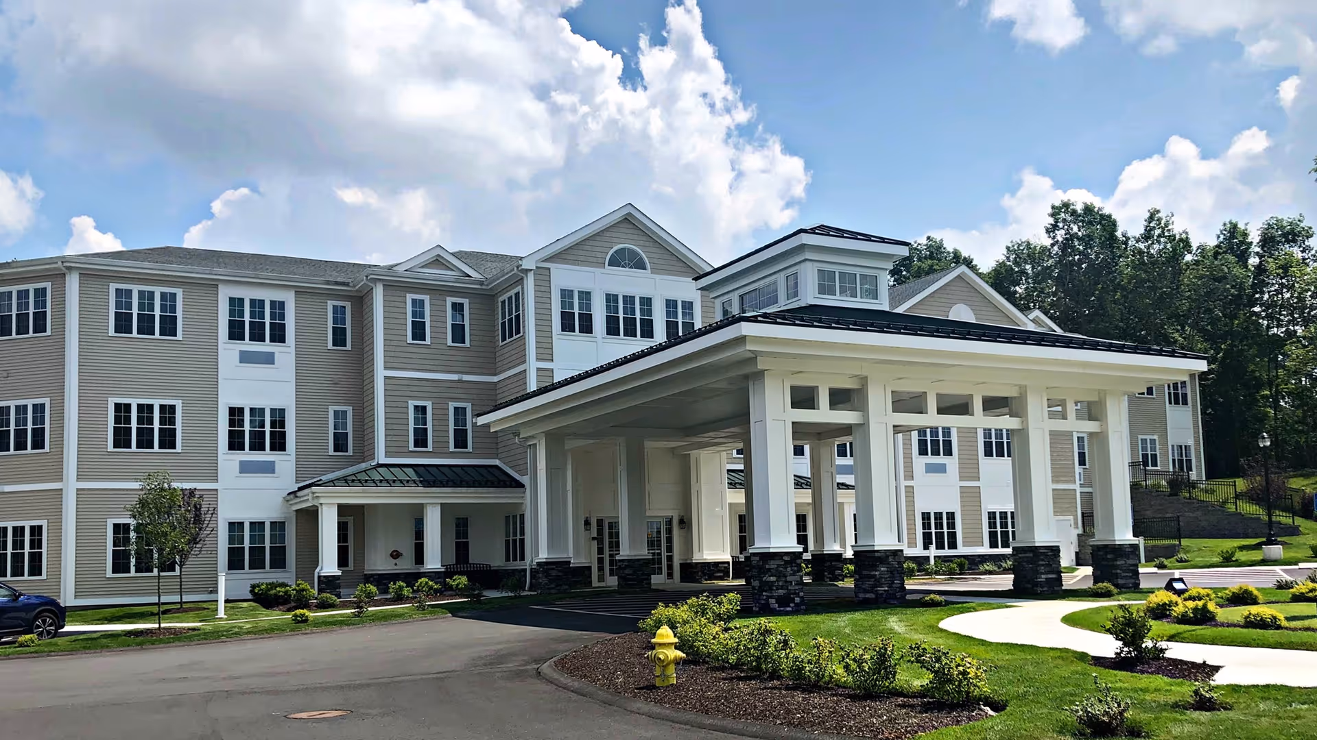 Exterior view of The Ivy at Watertown Assisted Living and Memory Care building on a sunny day with blue sky and some clouds. The building is three stories tall with beige siding and white trim. There is a large covered entrance supported by white columns with stone bases. Well-maintained landscaping with green grass, bushes, and a yellow fire hydrant is visible in front of the building.